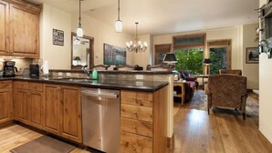 A spacious kitchen with wooden cabinets, a built-in dishwasher, and a large island. There is a chandelier hanging from the ceiling and a dining area with a table and chairs.