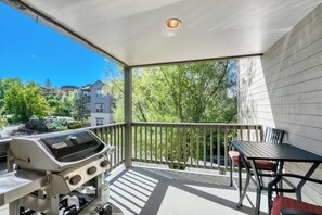 A balcony with a grill, a table, and chairs, overlooking a residential area with trees and buildings.