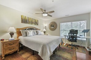 Ein Schlafzimmer mit Bett, Nachttischlampen, Schreibtisch mit Stuhl, Deckenventilator, Fenster mit Jalousien und einem Wandspiegel.