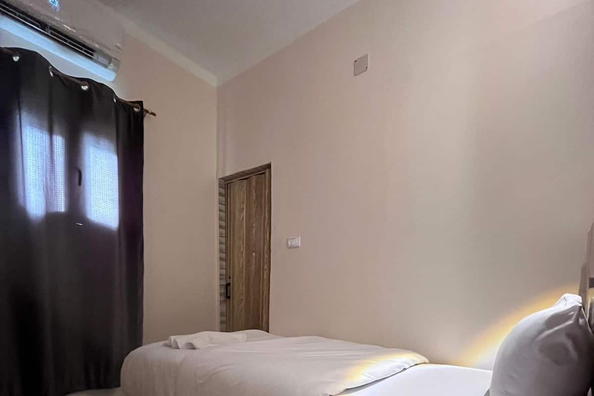 Uma cama de solteiro com colchão e almofadas brancas, uma mesa de cabeceira com controle remoto e uma janela com cortinas escuras.
