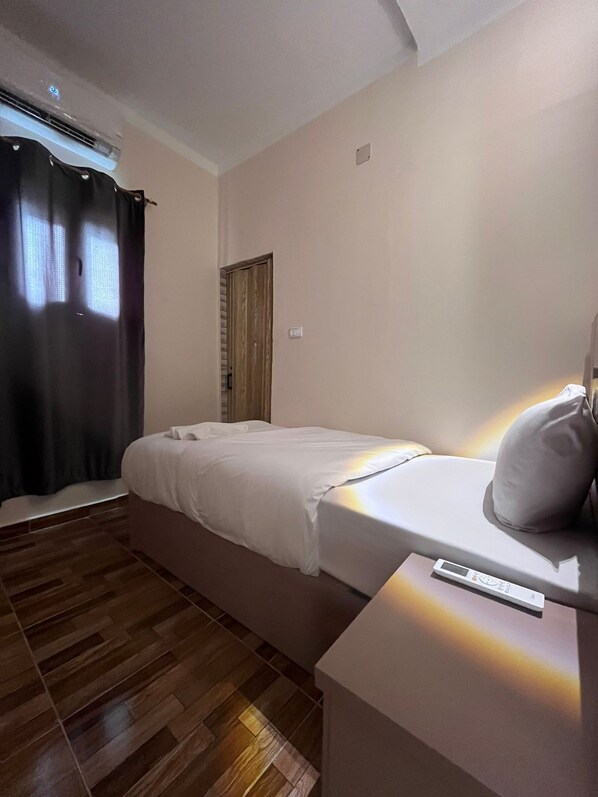 A single bed with a white mattress and pillows, a bedside table with a remote control, and a window with dark curtains. - Cairo casa inn (Cairo)