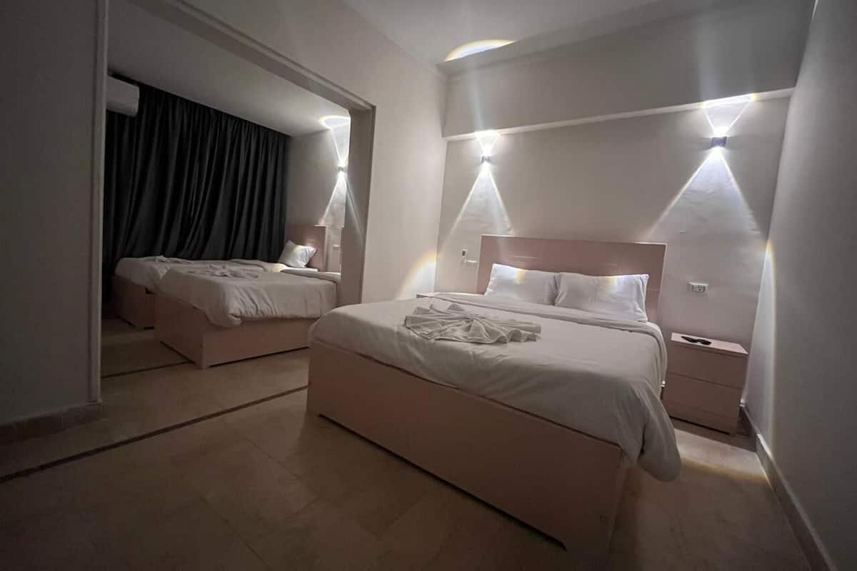 Quarto de hotel com uma cama grande, uma cama menor e uma mesa de cabeceira. Há duas luzes fixadas na parede e um espelho sobre a cama.