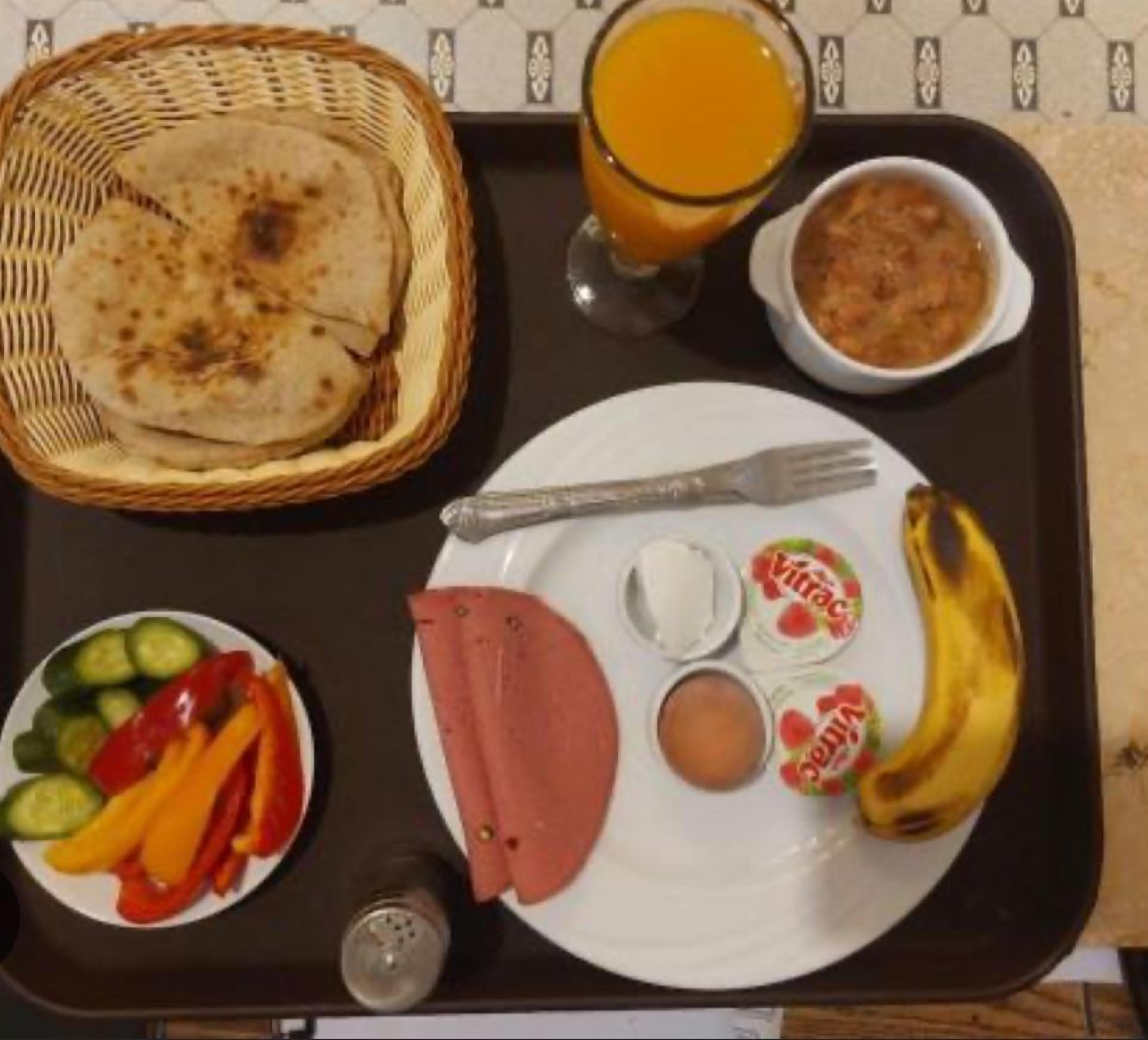 Bandeja de desayuno con tostadas, jugo de naranja, un tazón de sopa, un plato con huevos, verduras, jamón y un plátano.
