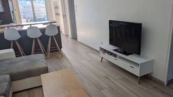 Un salon moderne comprenant un canapé, une table basse, un téléviseur posé sur un meuble blanc, et un coin cuisine avec un bar et des tabourets.