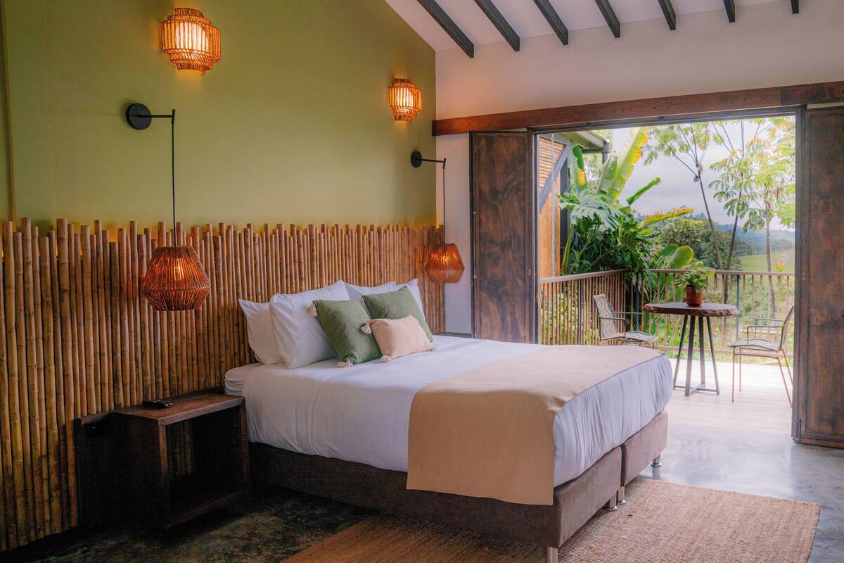 A bedroom with a bed, bedside tables, a balcony with a table and chairs, and a view of greenery.