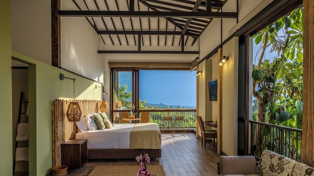 Un dormitorio con cama, sofá, una mesita, ventana con vistas y balcón con plantas.