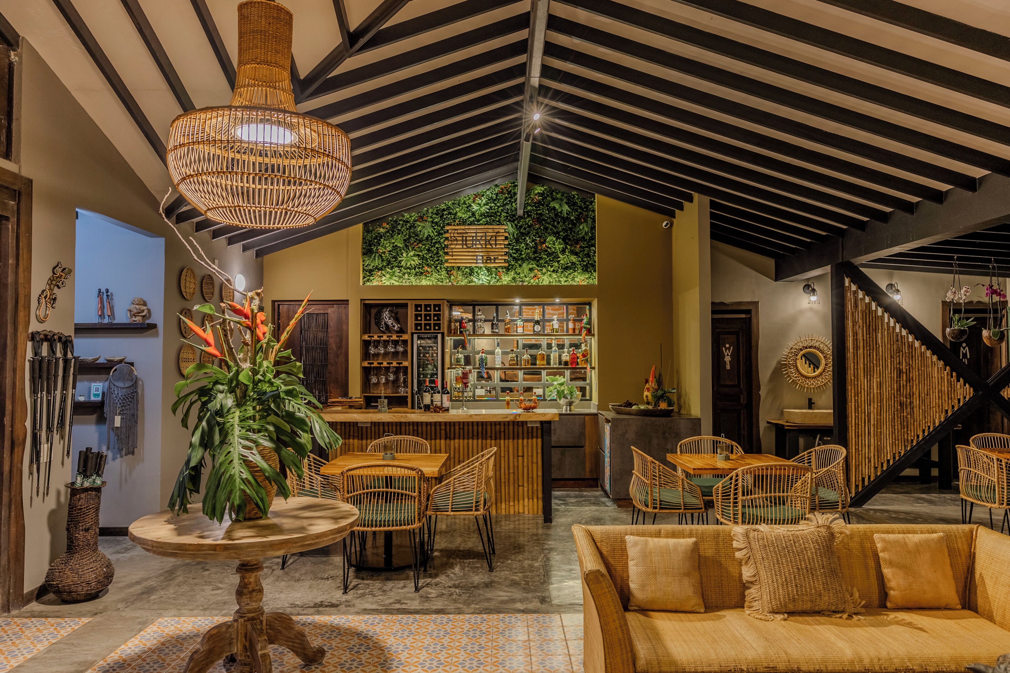 A bar area with a wooden counter and wicker chairs, a round wooden table with chairs, and a staircase with a black railing.