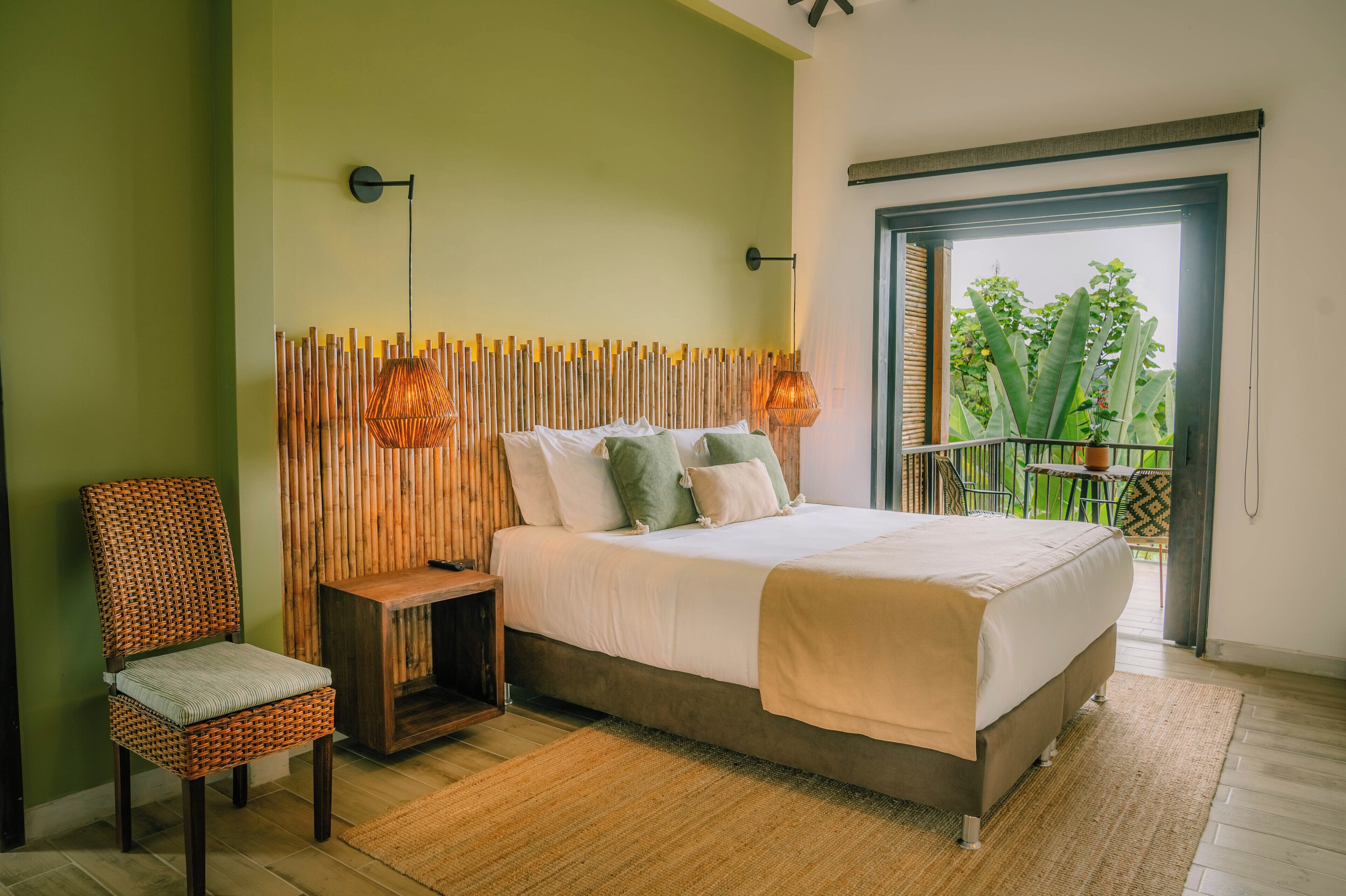 A bedroom with a bed, a chair, a nightstand, a wall-mounted lamp, and a view of greenery through an open door.