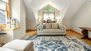 Una camera da letto mansardata accogliente, con soffitto spiovente, una grande finestra, un divano, un tappeto decorato e una panca in legno.