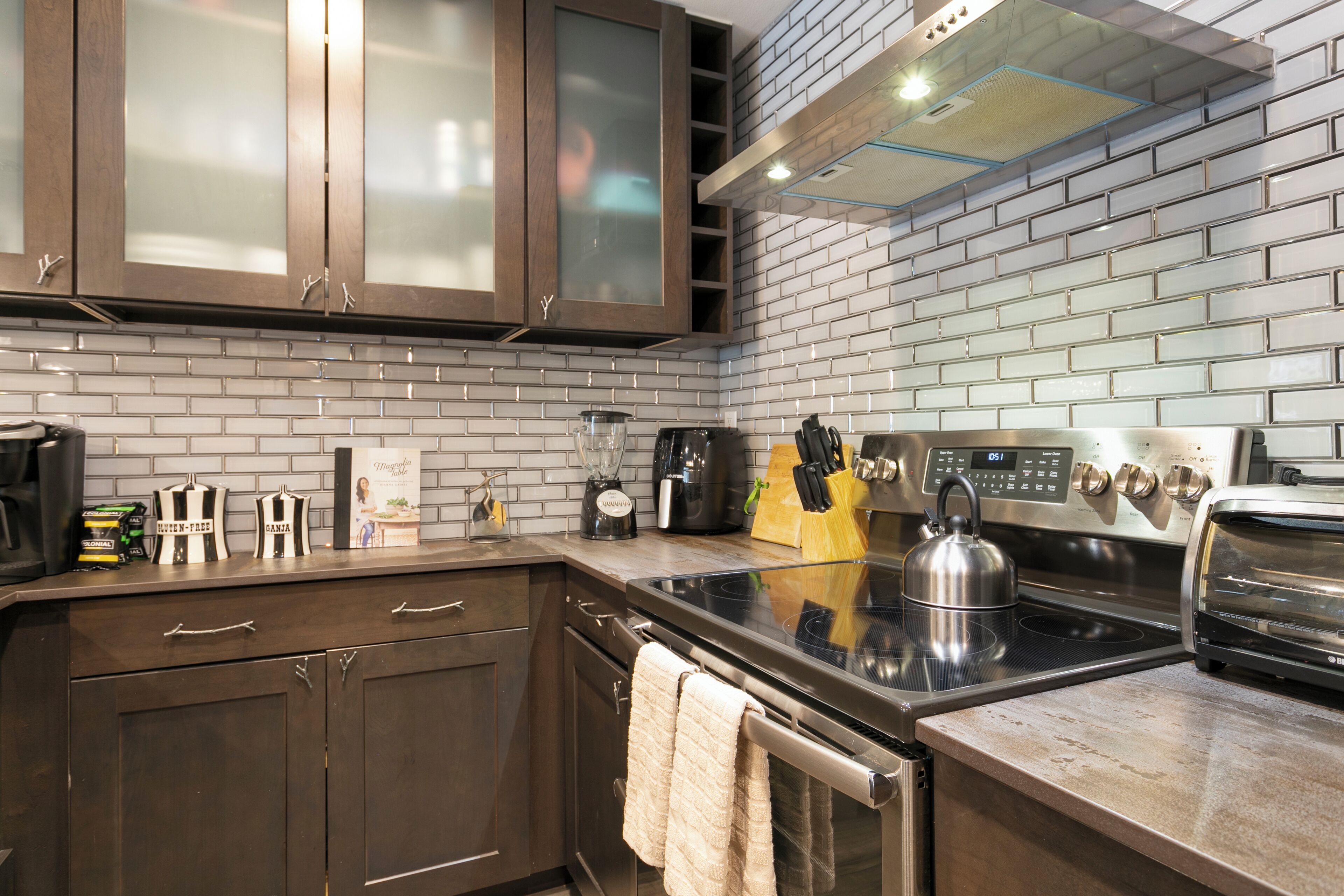 A modern kitchen with dark wooden cabinets, a brick backsplash, and stainless steel appliances including a kettle, toaster, and coffee maker.