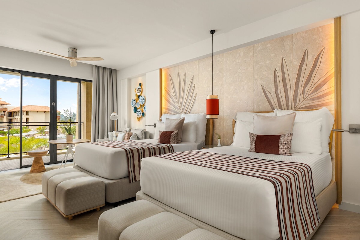A modern hotel room with a large bed, a sofa, a dining table, and a view of the outdoors.