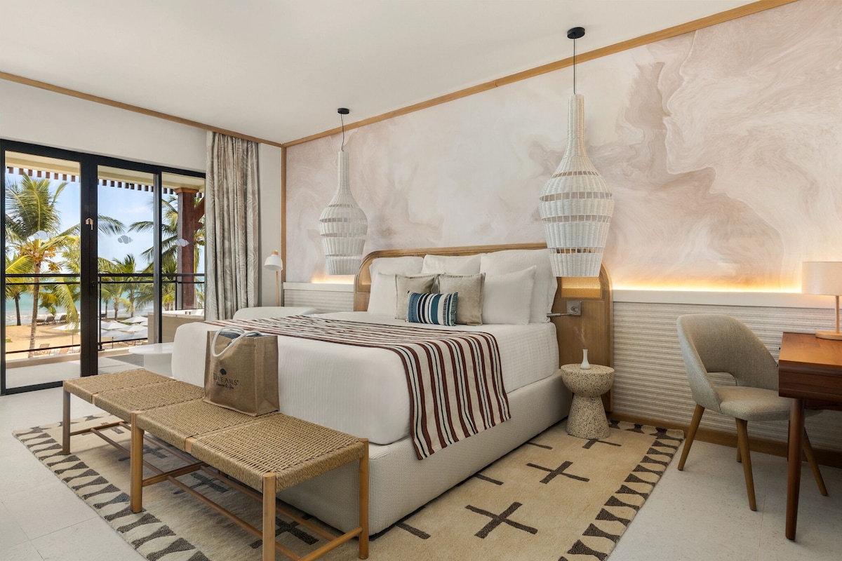 A modern bedroom with a bed, bedside table, chair, and a view of palm trees.