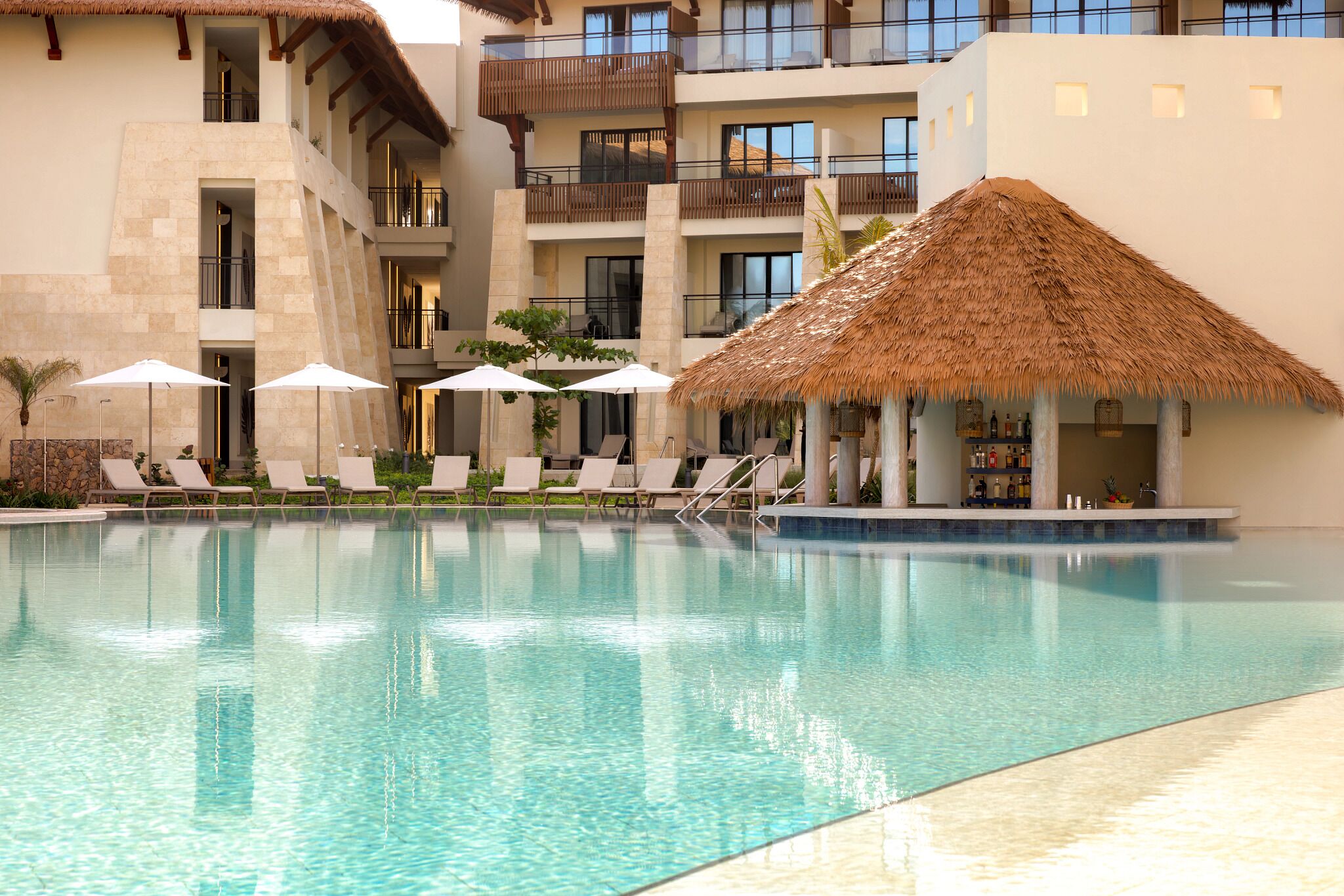 Ein Hotelpool mit Liegestühlen und Sonnenschirmen, umgeben von einem mehrstöckigen Gebäude mit Balkonen.