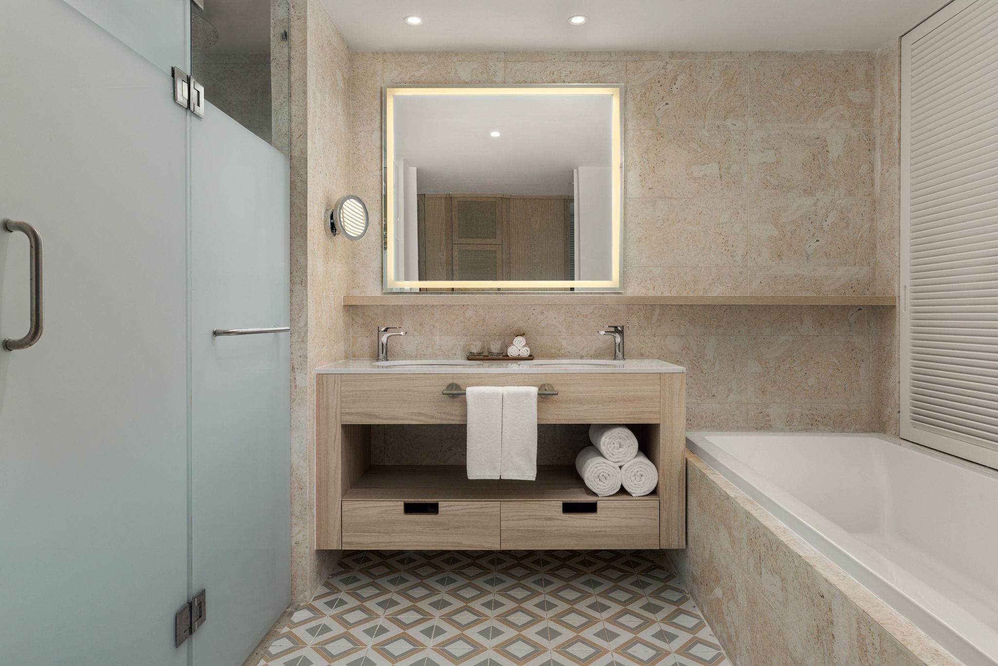 A modern bathroom with a marble wall, a large mirror, a wooden vanity, and a bathtub.
