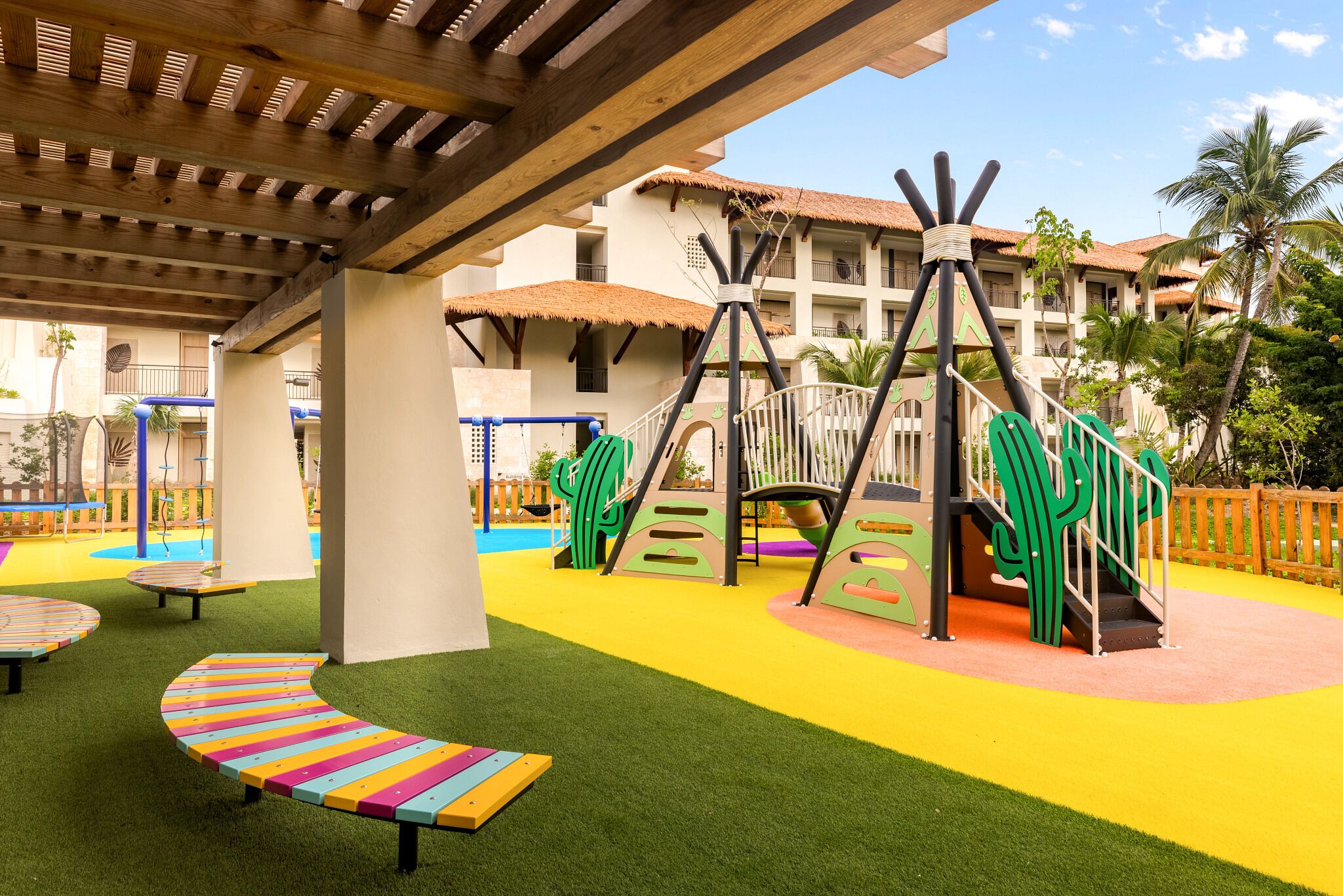 A playground with colorful play structures, artificial grass, and a building with a thatched roof in the background.