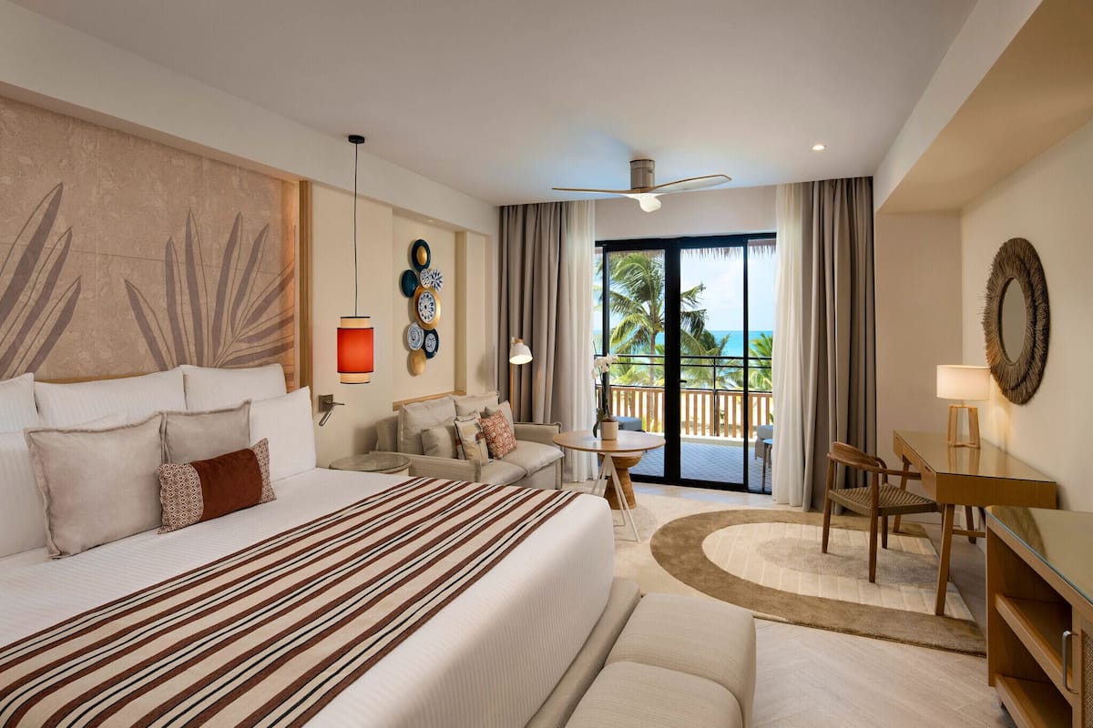 A hotel room with a large bed, a desk, a chair, a sofa, a balcony with a view, and a ceiling fan.