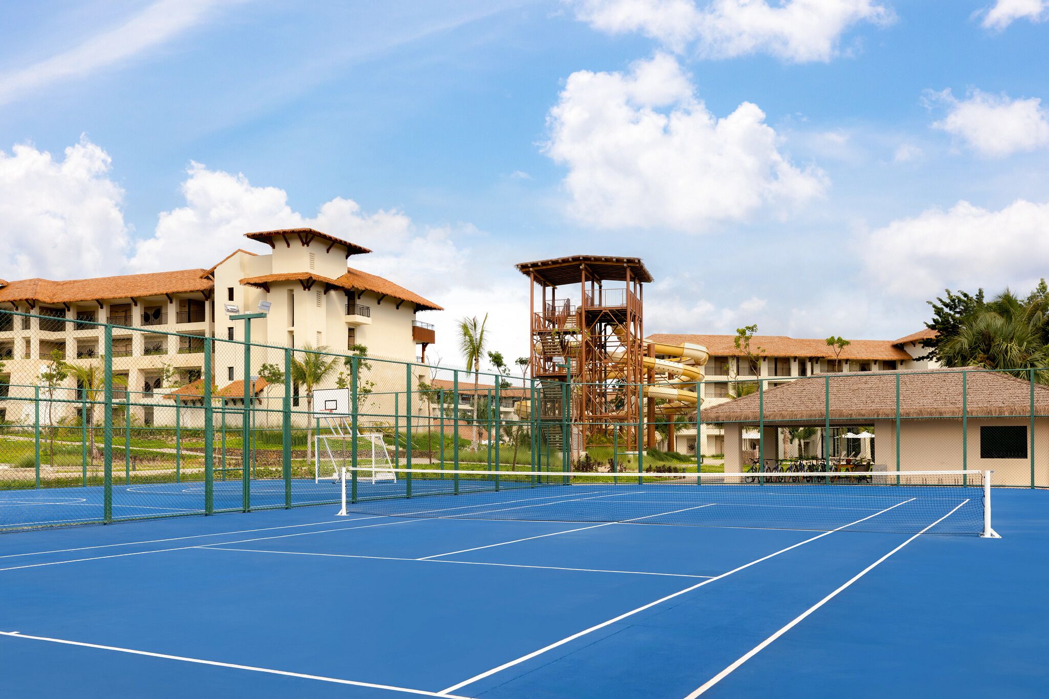 A tennis court with a net, surrounded by a fence, and buildings in the background.