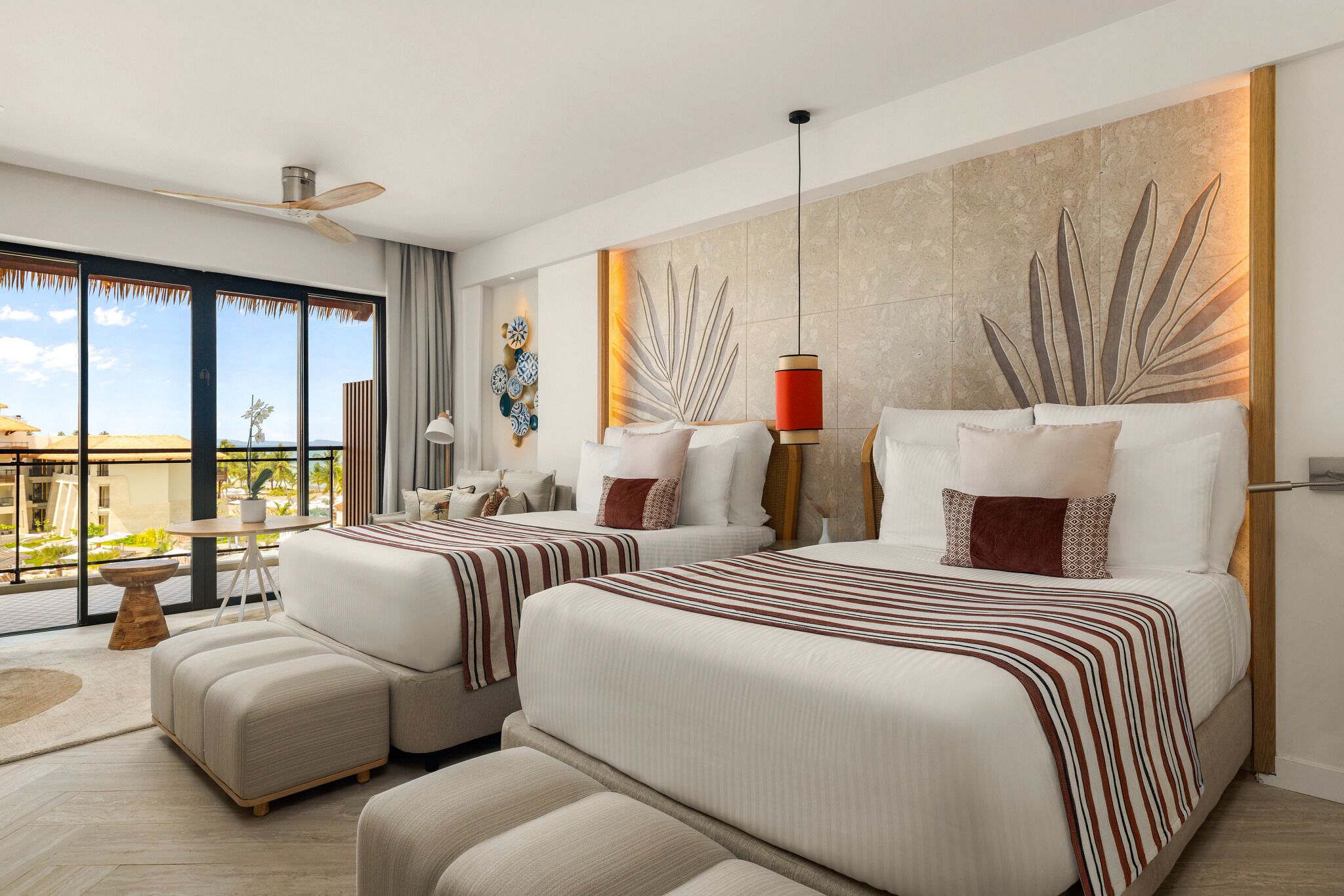 A modern hotel room with a large bed, two bedside tables, a seating area with a sofa and armchairs, a dining table, and a balcony with a view.