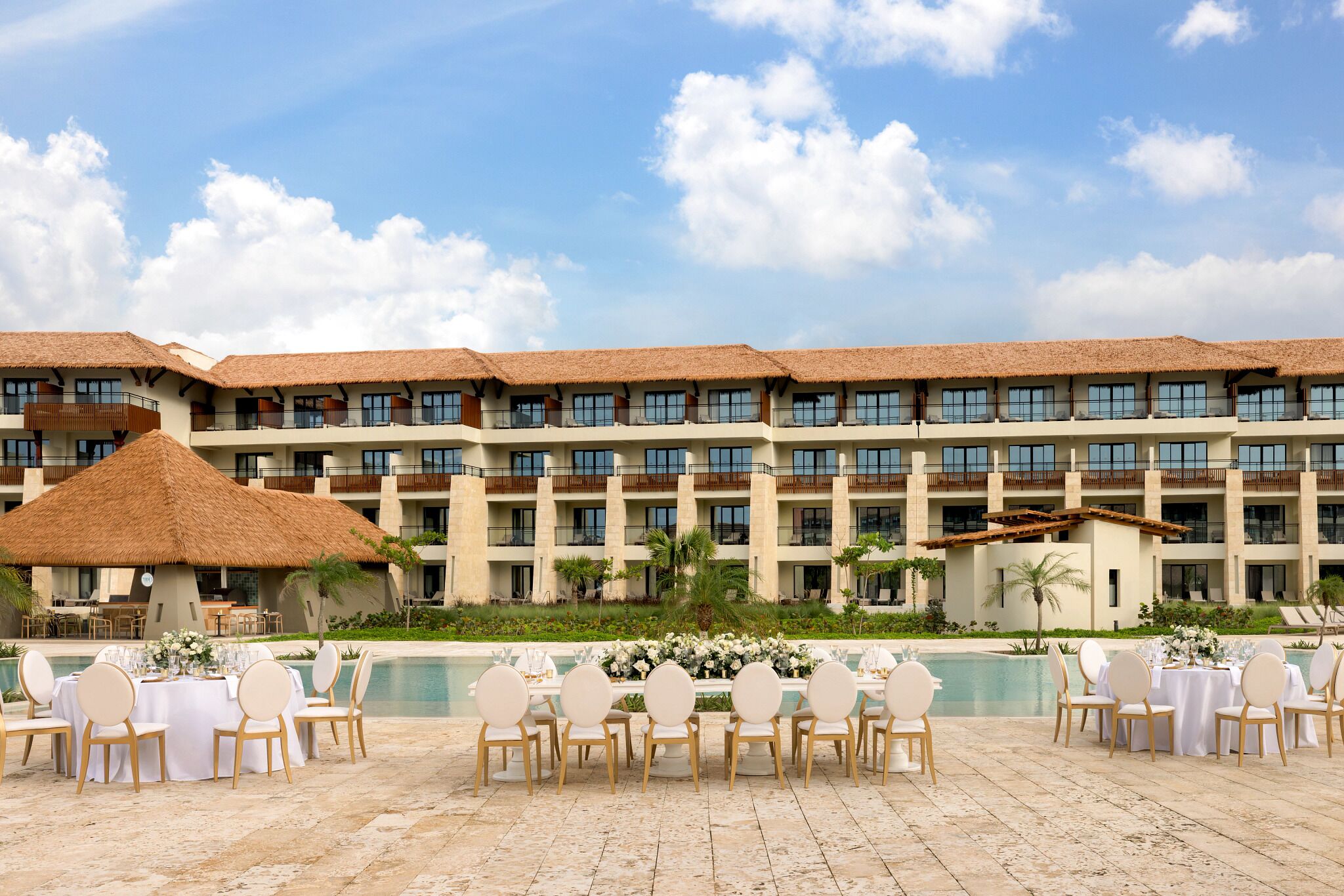 A hotel with multiple balconies, a pool area with tables and chairs, and a thatched-roof structure.