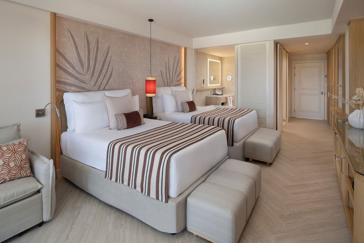 A modern hotel room with two beds, a sitting area, a vanity with a mirror, and a wardrobe.
