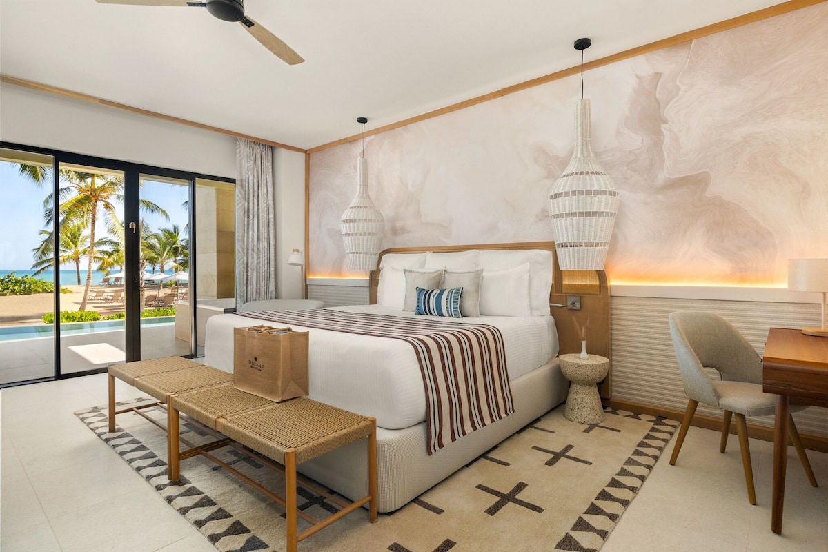 A modern hotel bedroom with a large bed, a desk, a chair, a bench, and a view of the beach.