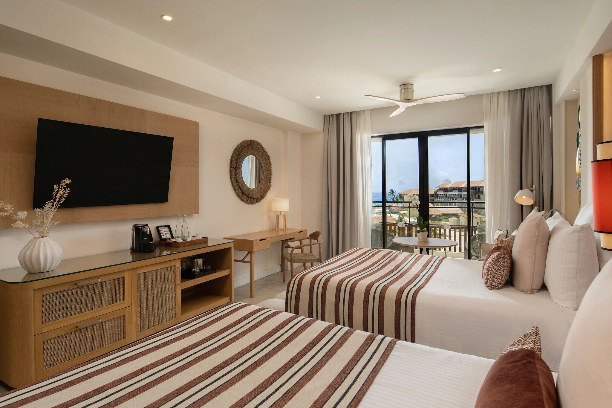 A hotel room with two beds, a desk, a TV, a balcony with a view, and a ceiling fan.