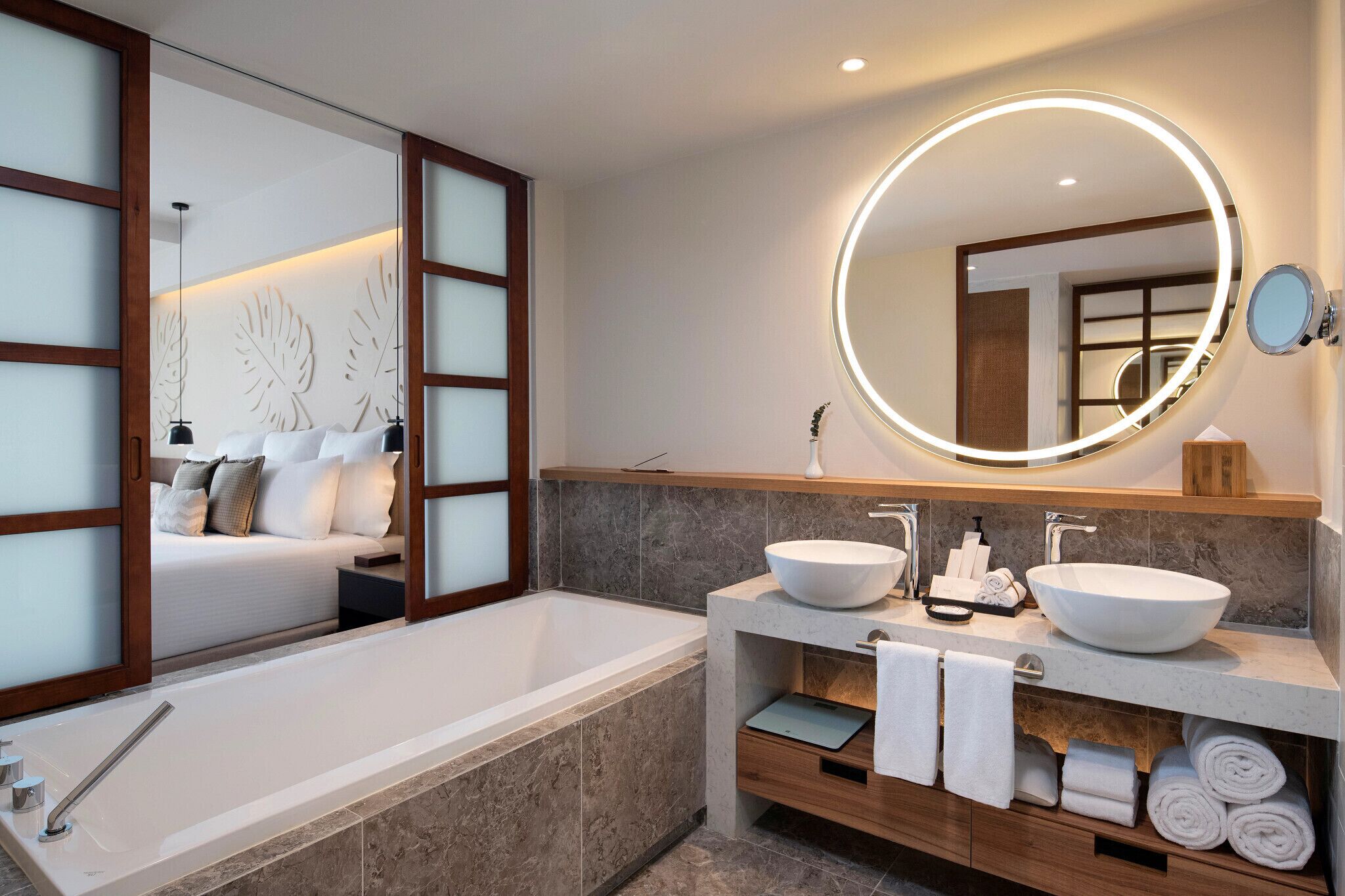 A modern bathroom with a large bathtub, two vessel sinks, a round mirror with lighting, and a wooden vanity with towels.