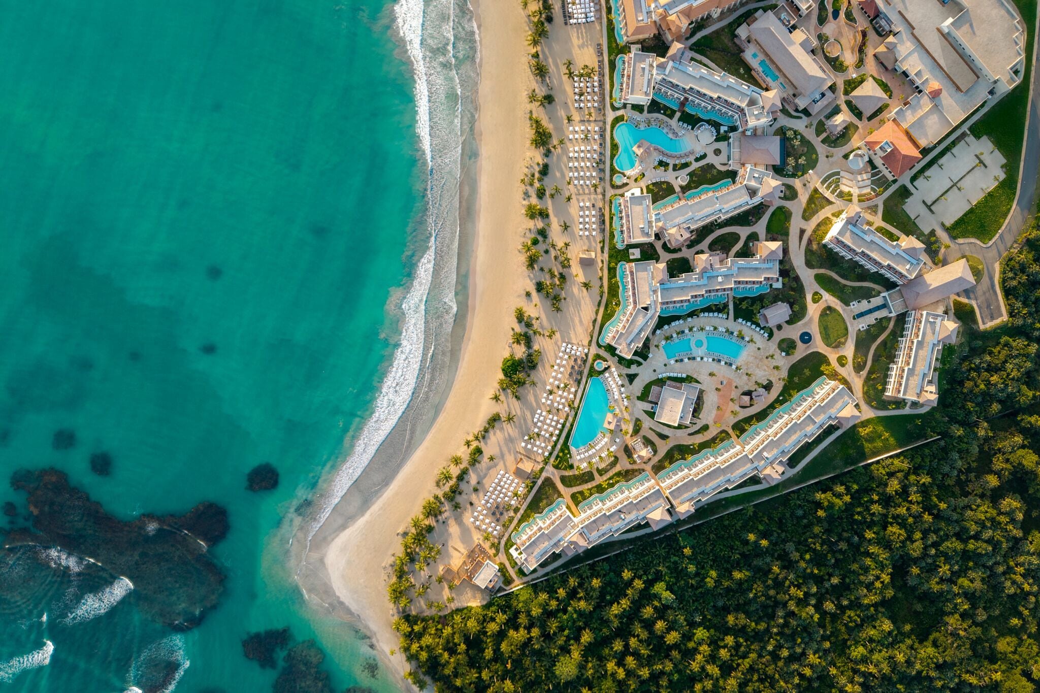 An aerial view of a resort with a beachfront, multiple swimming pools, and a variety of building structures.