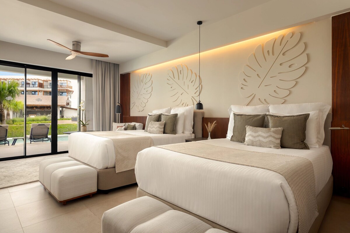 A hotel room with two beds, a ceiling fan, a large window with a view of a building and greenery, and a wall-mounted leaf decoration.