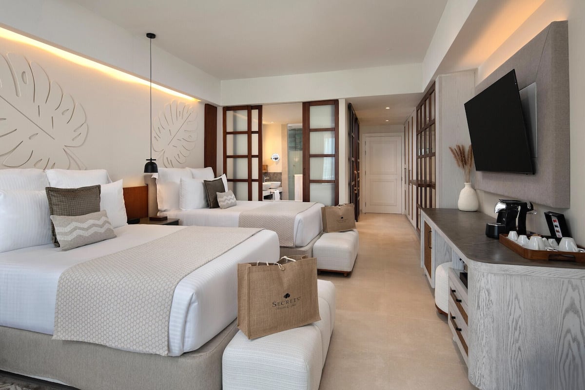 A hotel room with two beds, a minibar, a TV, and a large leaf-patterned wall.