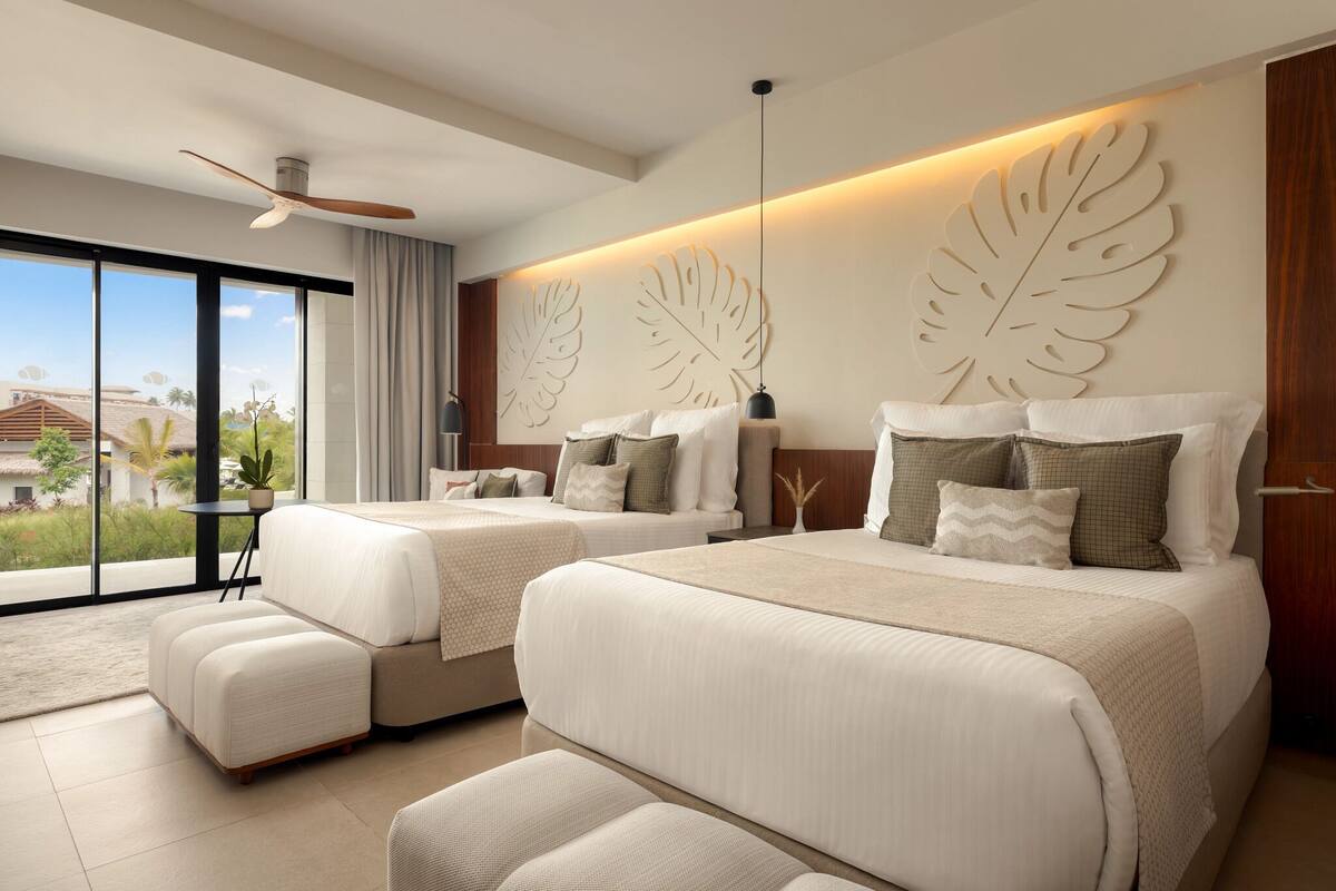 A modern hotel room with two beds, a ceiling fan, large windows, and a view of the outdoors.
