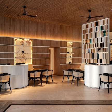 A hotel lobby with wooden paneling, a reception desk, two seating areas with tables and chairs, and a ceiling fan.