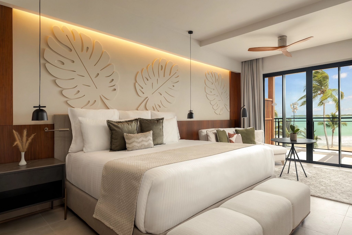 A modern hotel room with a large bed, a ceiling fan, a view of the ocean, and a small table with a plant.