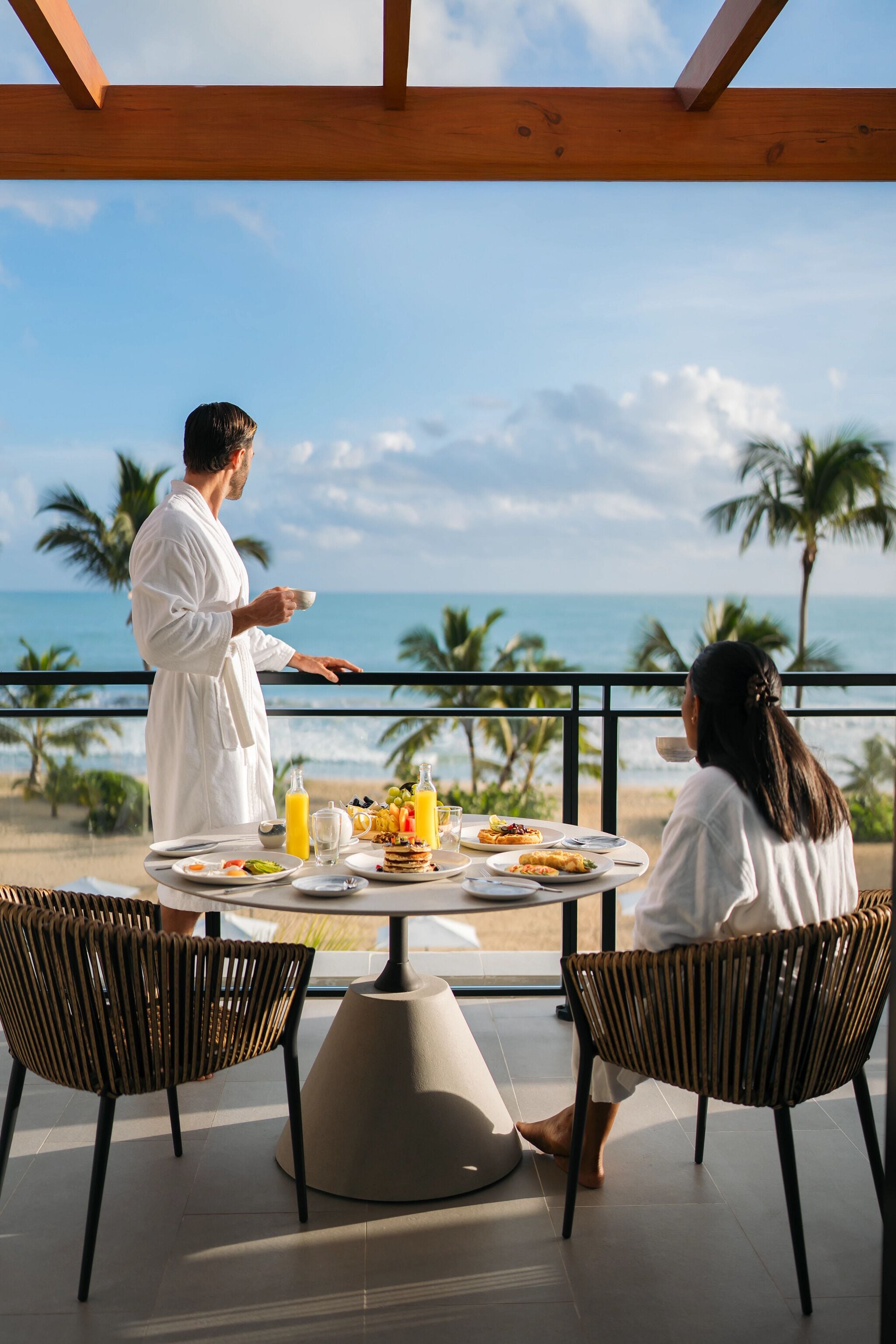 A dining table with breakfast items, two wicker chairs, and a view of the ocean.