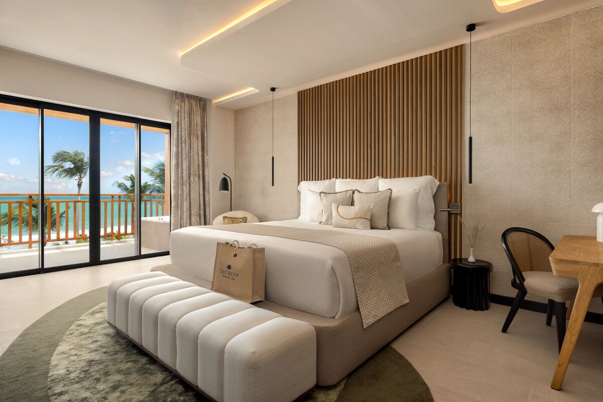 A modern hotel room with a large bed, a seating area, a wooden accent wall, and a view of the ocean.