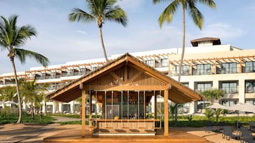 A beachside bar with a wooden structure, palm trees, and a hotel in the background.