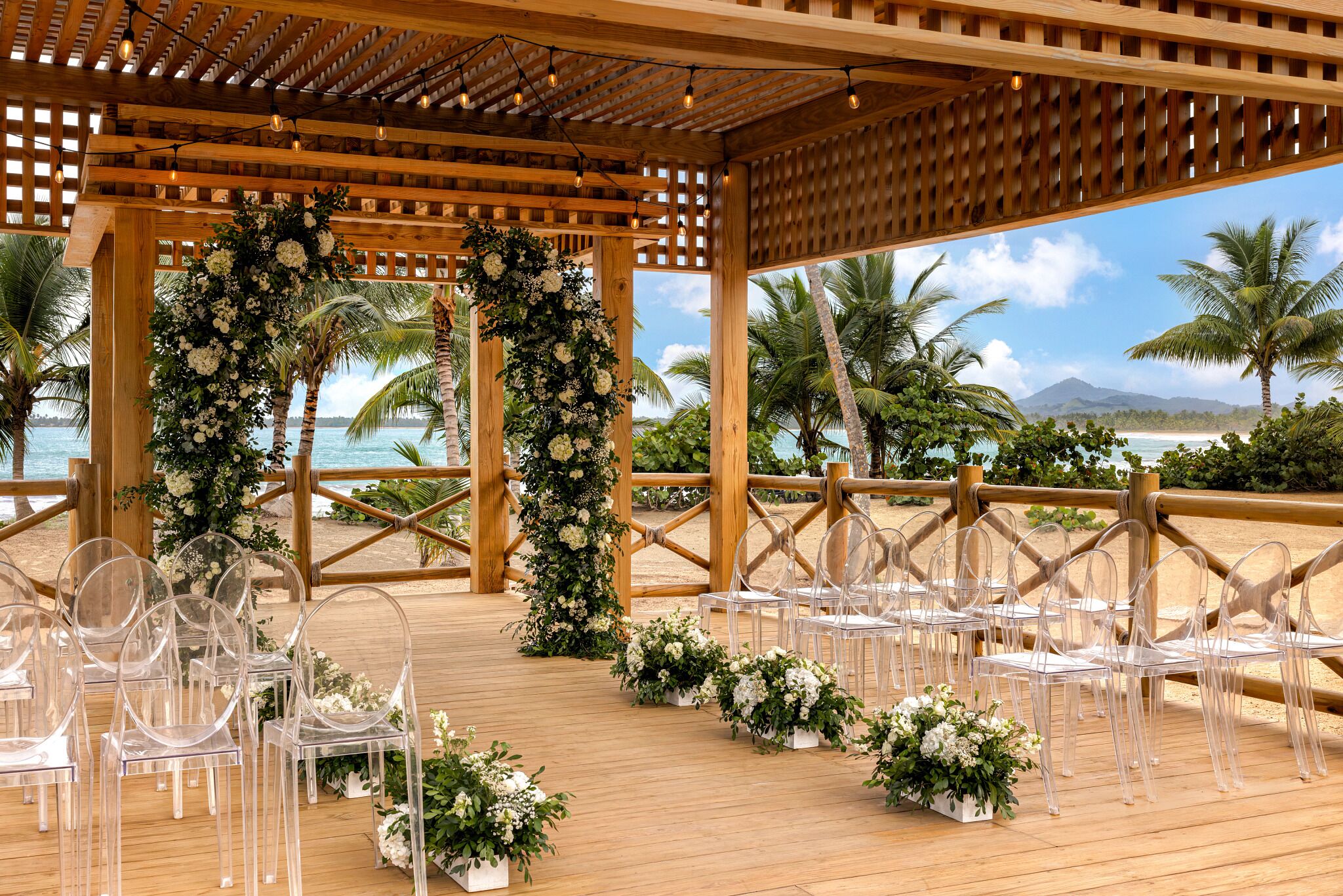 A wooden deck with clear chairs and floral arrangements, overlooking a beach with palm trees and a clear sky.