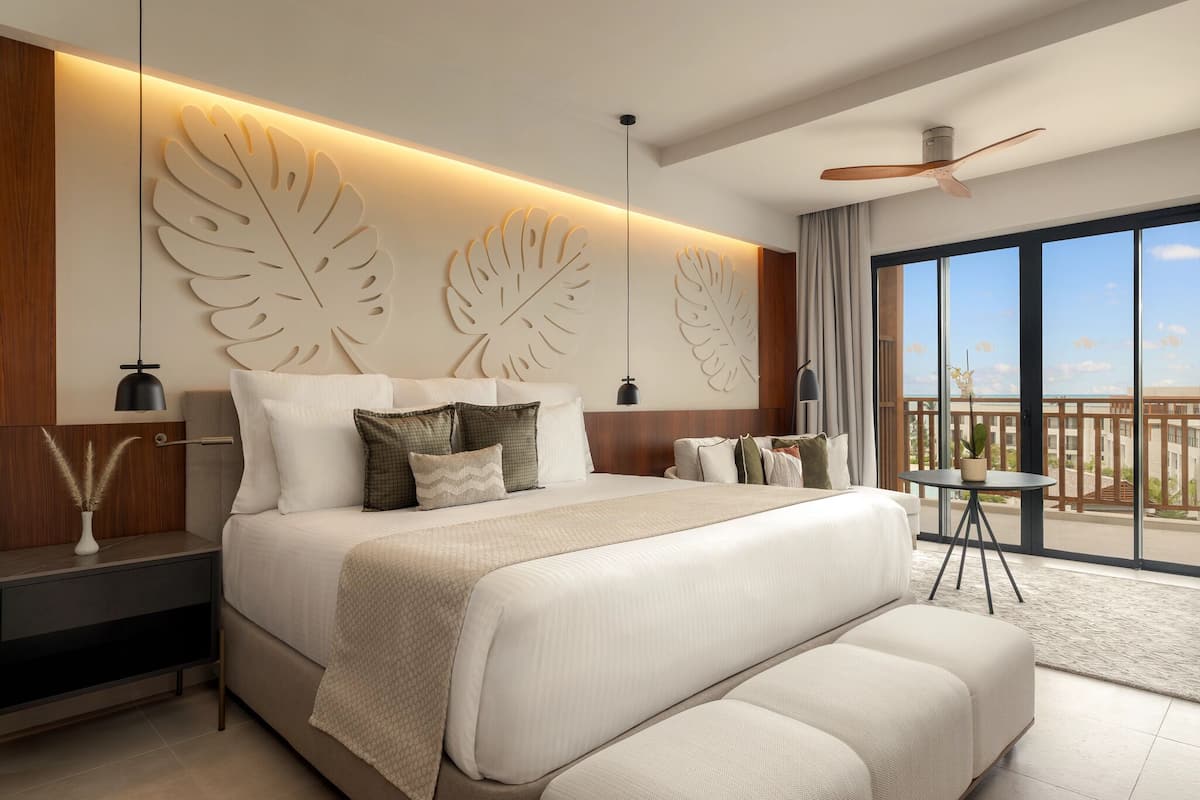 A modern hotel room with a large bed, a ceiling fan, a balcony with a view, and a nightstand with a lamp.