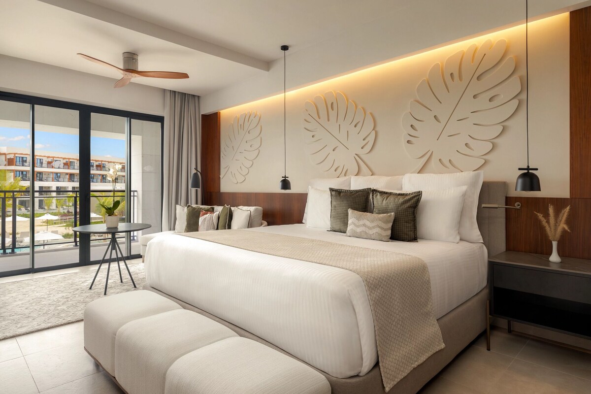 A modern bedroom with a large bed, a ceiling fan, a view of the outside, and a nightstand with a lamp.