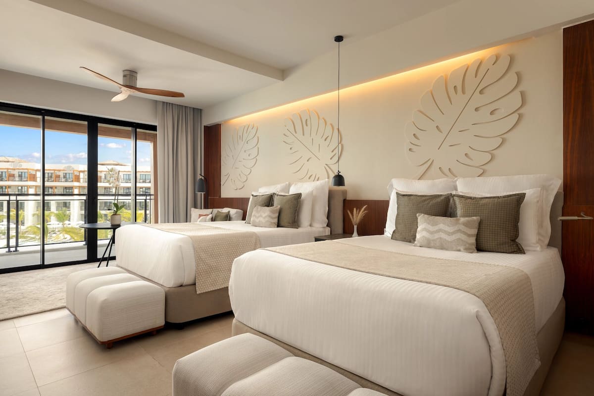 A modern hotel room with two beds, a ceiling fan, large windows, and a view of buildings.