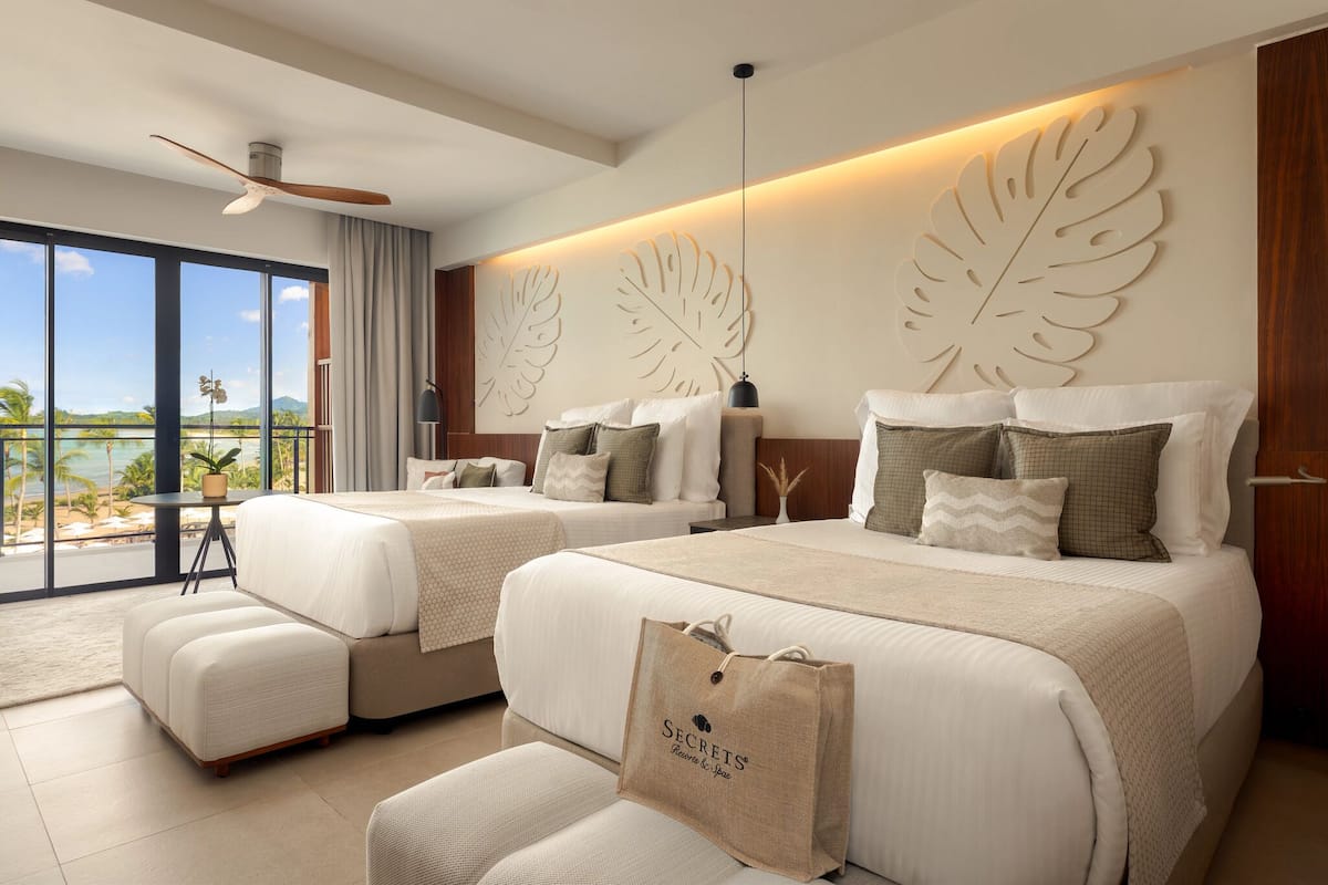 A hotel room with two beds, a balcony with a view, a ceiling fan, and a large leaf wall decoration.