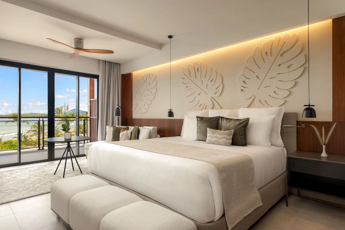 A modern bedroom with a large bed, a balcony with a view, and a ceiling fan.
