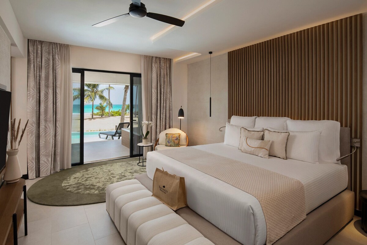 A modern hotel room with a large bed, a ceiling fan, a view of the beach, and a small table with a lamp.