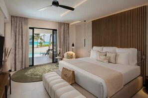 A modern hotel room with a large bed, a ceiling fan, a view of the beach, and a small table with a lamp.