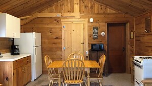 A cabin-style kitchen with wooden walls, a dining table with four chairs, a refrigerator, a microwave, and a stove. - 1 Bedroom Riverside Cabin in The Forks, ME (The Forks)