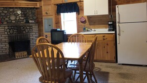 A wooden cabin interior with a stone fireplace, wooden dining table, chairs, a kitchen area with a refrigerator, and a window with curtains. - 1 Bedroom Riverside Cabin in The Forks, ME (The Forks)