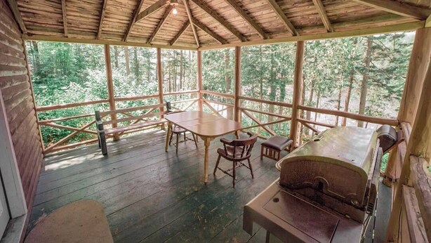 A wooden deck with a table and chairs, surrounded by a wooden railing, overlooking a forested area. - 1 Bedroom Riverside Cabin in The Forks, ME (The Forks)