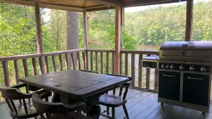 A covered outdoor area with a wooden table and chairs, a grill, and a view of a forested area. - Riverside Cabin in The Forks (The Forks)