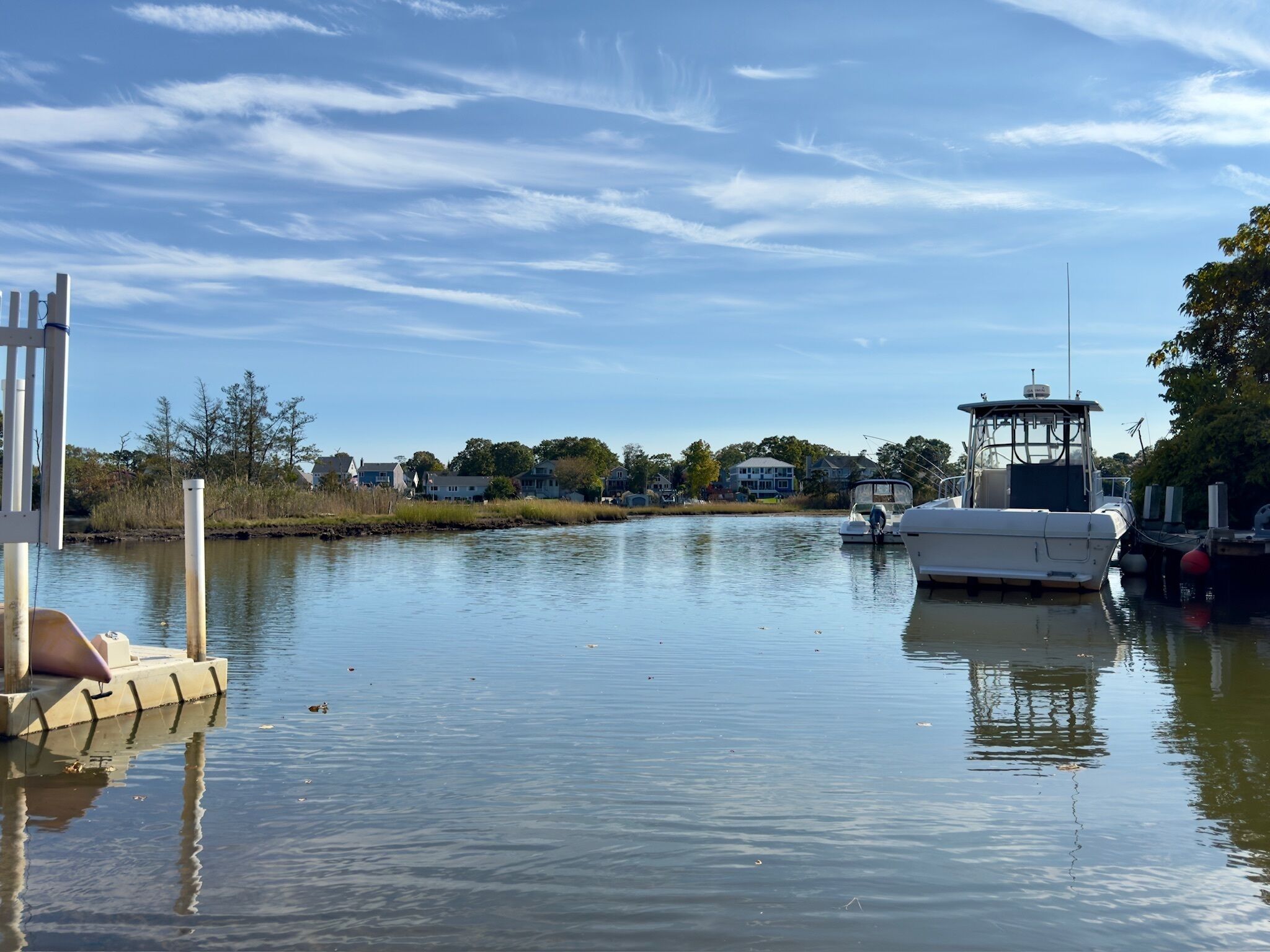 A calm waterway with boats docked, a pier, and houses in the background.
