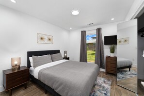 A modern bedroom with a large bed, a nightstand with a lamp, a dresser, and a TV on the wall. - The Gateway 6BED 2 Houses GameRoom Pool (Cedar Park)